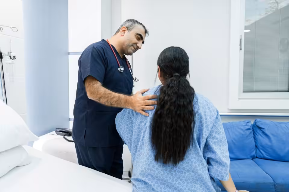A healthcare professional with a stethoscope speaks to a patient in a hospital gown, gently placing a hand on her shoulder in a clinical setting.