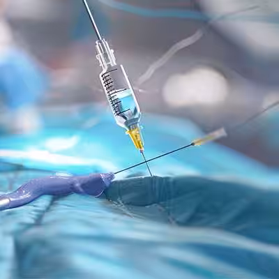 A syringe with a yellow-tipped needle is inserted into a patient's skin in a clinical setting with medical equipment and blue sterile fabric visible.
