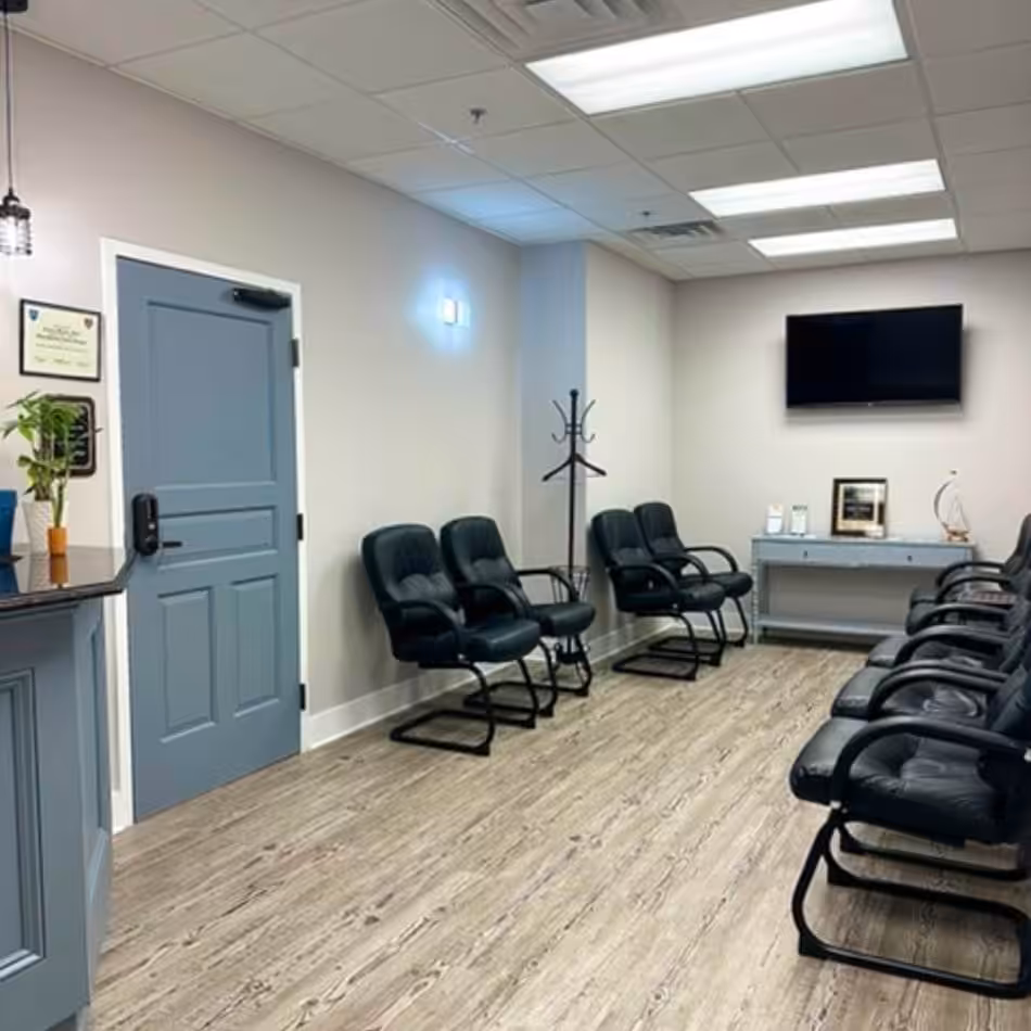 A waiting room with black chairs, a television, a coat rack, and framed certificates on a gray wall and door. The room has wood flooring and recessed ceiling lights.