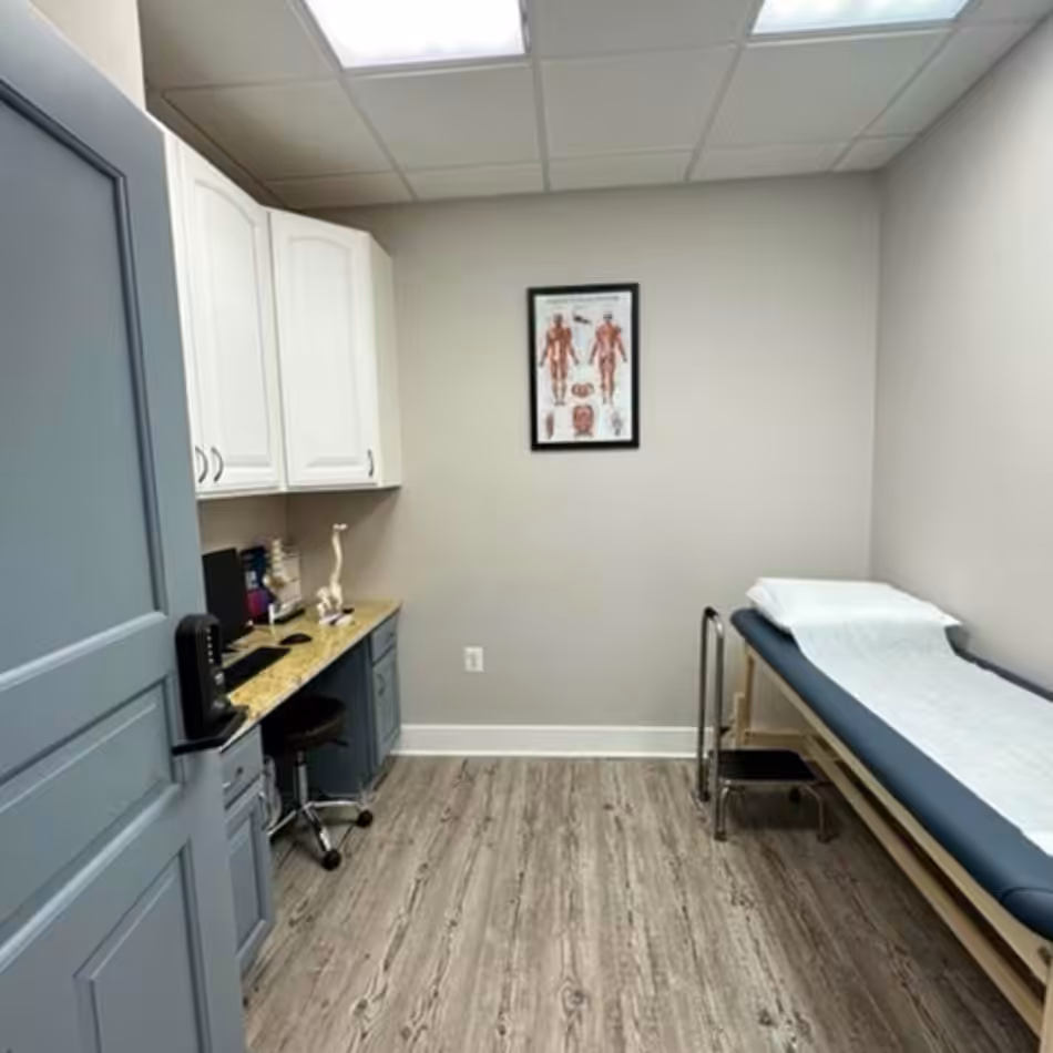 A small medical examination room with a treatment table, anatomical chart, cabinets, office chair, and a counter with medical supplies.