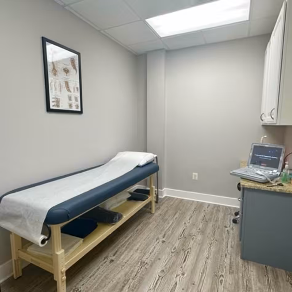 A small medical examination room with an examination table, wall art, an ultrasound machine on a counter, and wooden flooring.