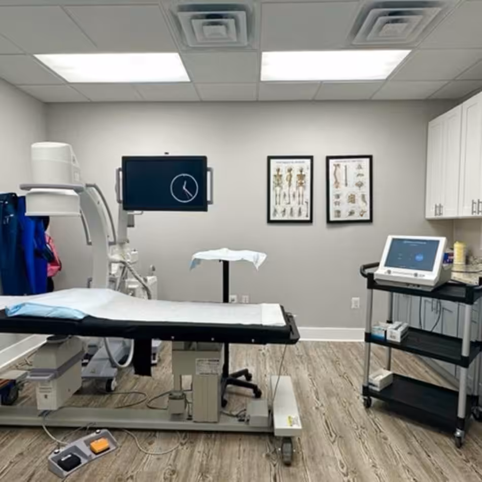 A medical examination room with a procedure table, medical equipment, wall-mounted monitor, anatomical charts, and shelves. The room appears well-lit and organized.