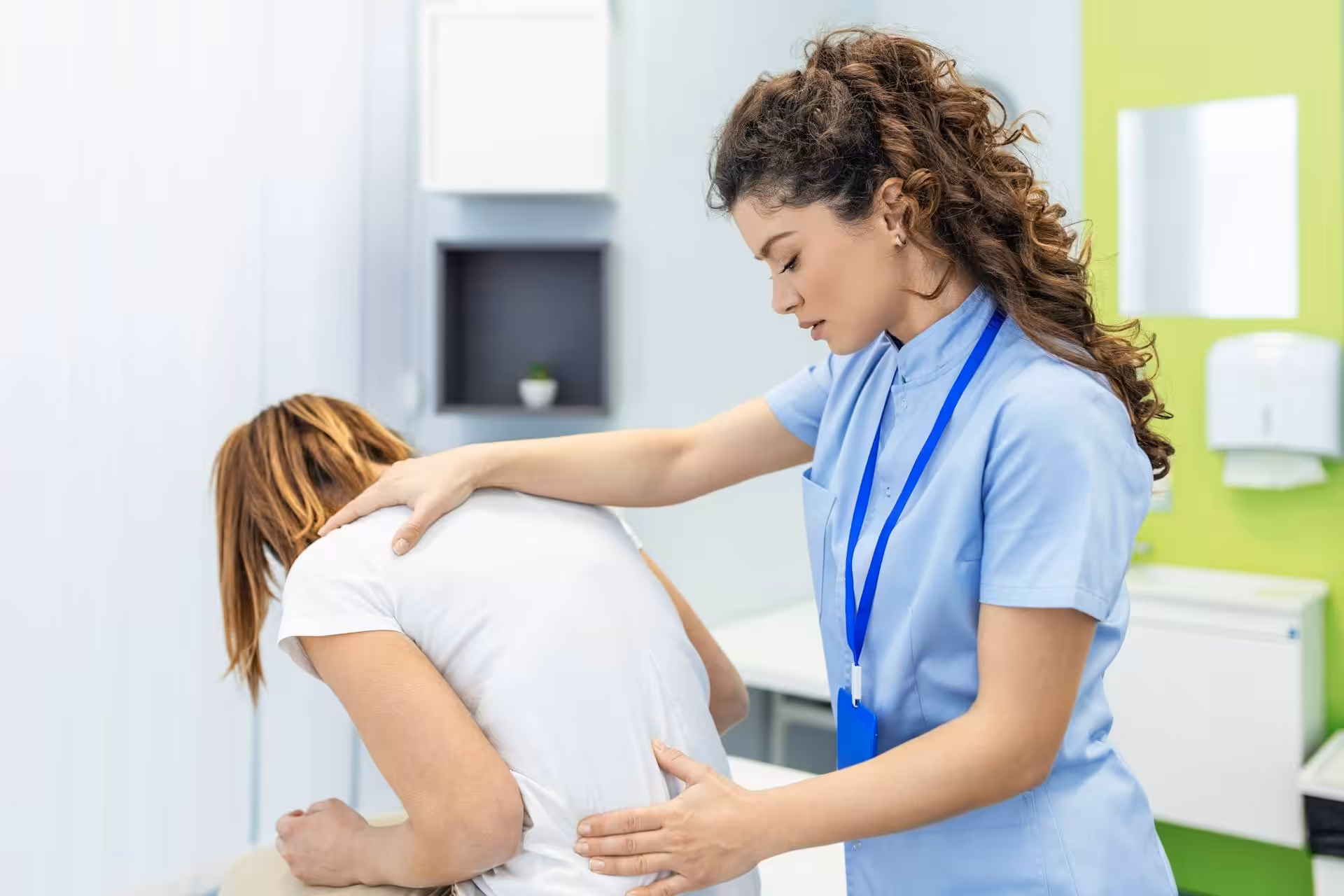 A healthcare professional at a pain clinic examines a patient’s back by gently pressing on their shoulder and lower back in a medical office.