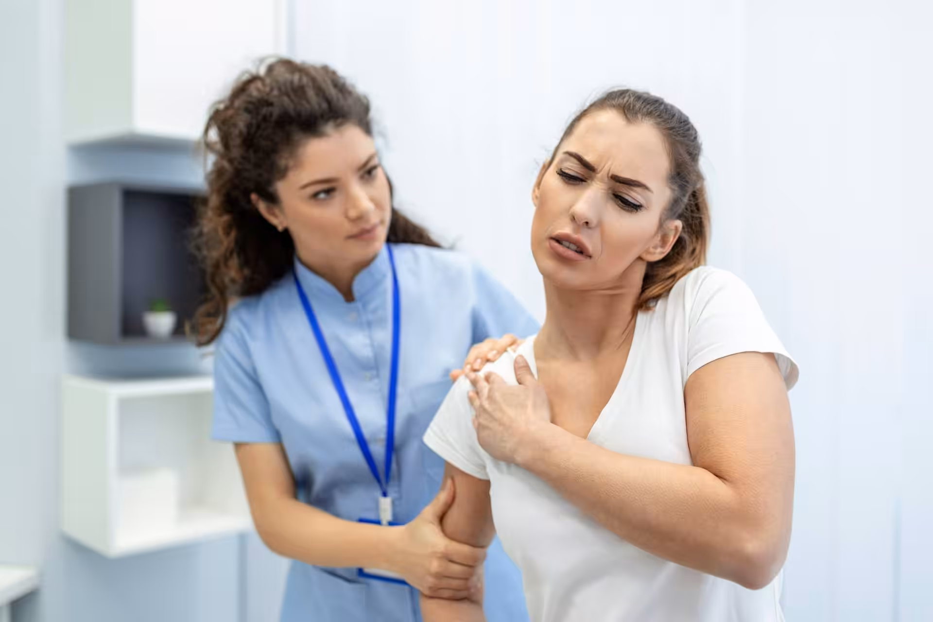 A healthcare professional at a pain clinic examines a woman who appears to be experiencing shoulder pain in a medical office setting.
