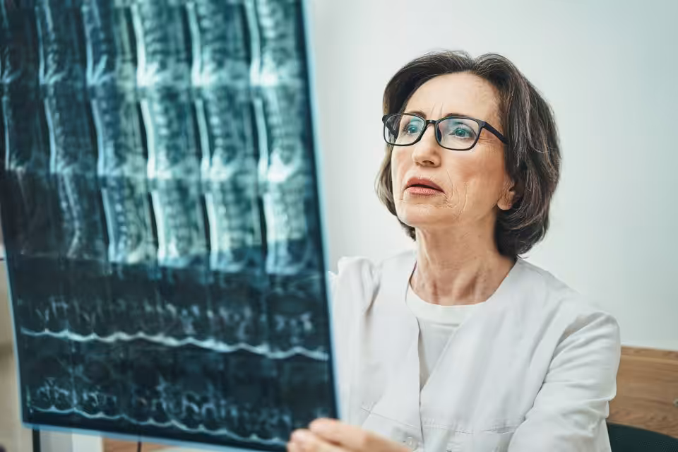 A woman in a white coat and glasses examines a spinal X-ray image, holding it up and looking at it closely.
