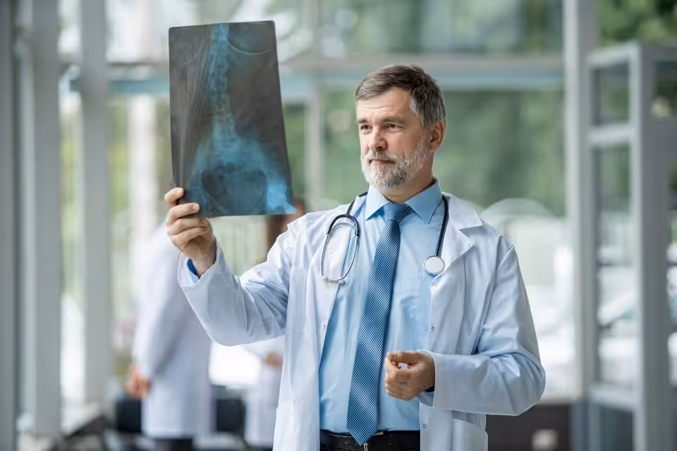 A doctor in a white coat and tie examines a spinal X-ray in a bright, modern medical facility.