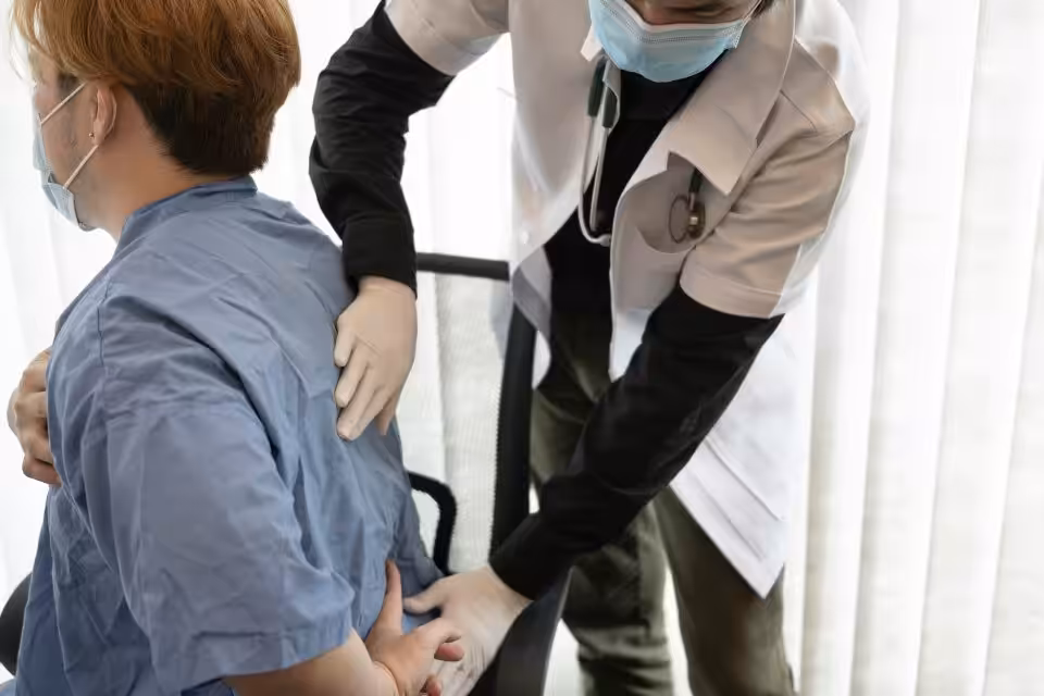 A healthcare professional in gloves examines a patient’s lower back. Both are wearing face masks; the patient is seated and wearing a blue medical gown.