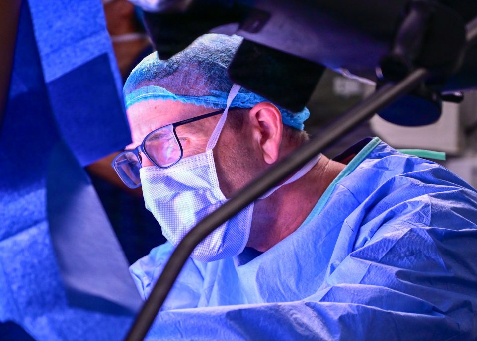 A surgeon in blue scrubs, a mask, and a hair cover focuses on a procedure in an operating room, with medical equipment visible in the foreground.