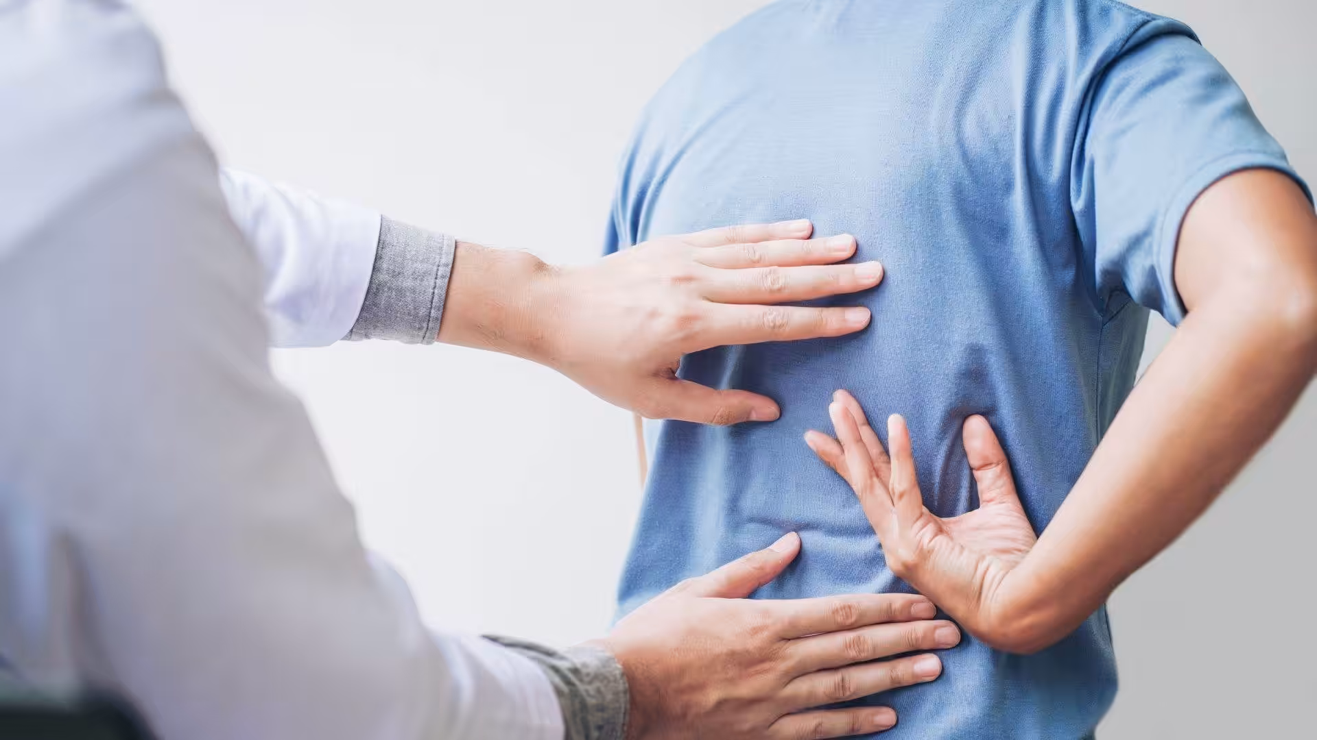 A person wearing a blue shirt holds their lower back in pain while a doctor in a white coat examines their back.